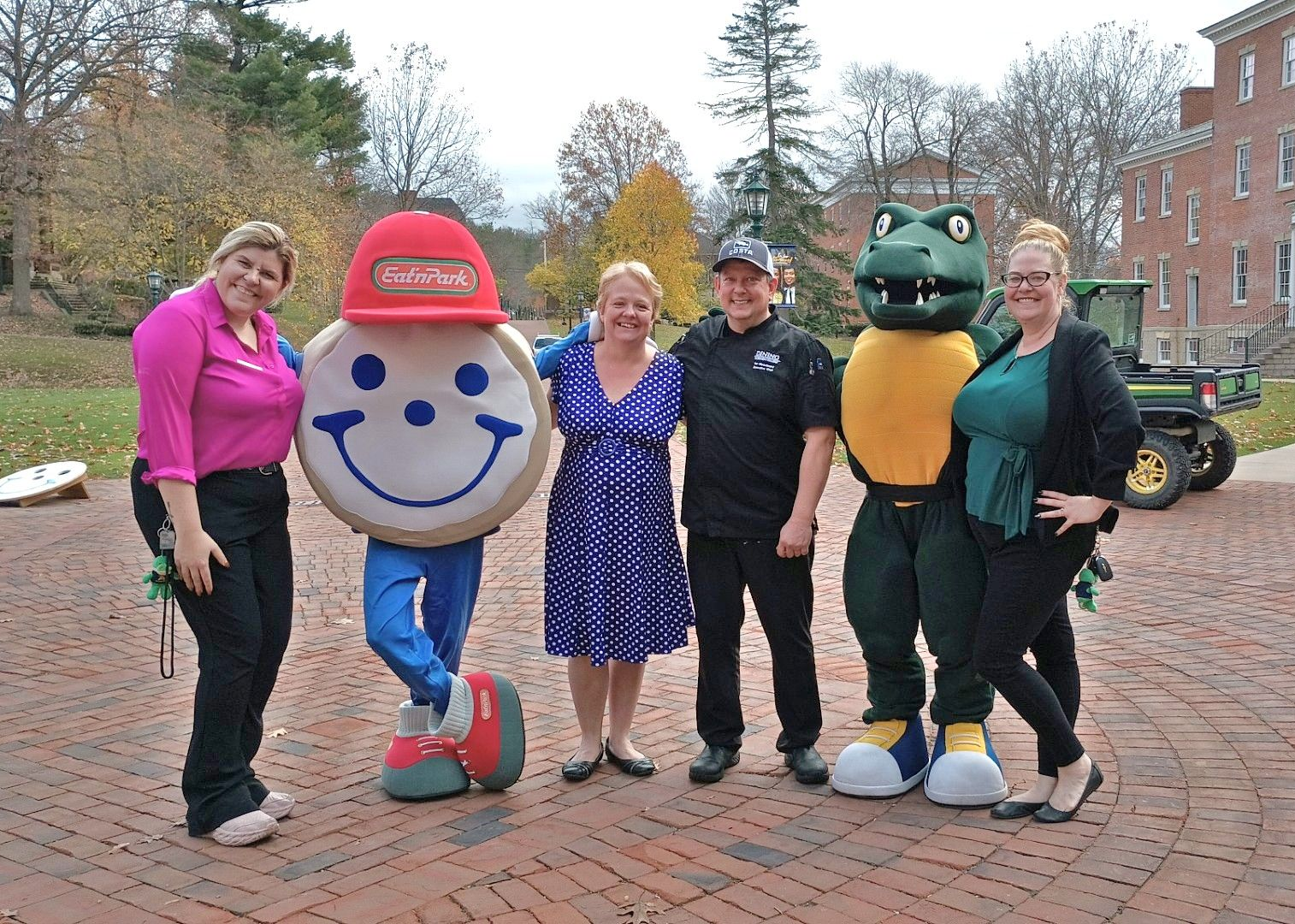 Group of people and mascots pose outdoors on a brick path in front of a brick building.