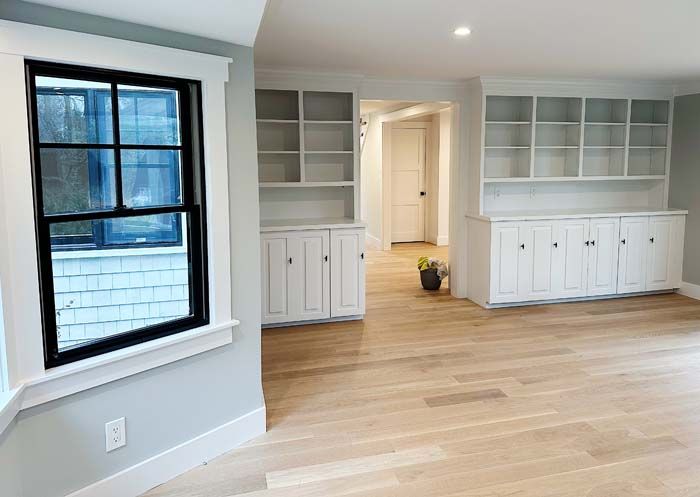 A living room with hardwood floors, white cabinets, shelves ,and a window.