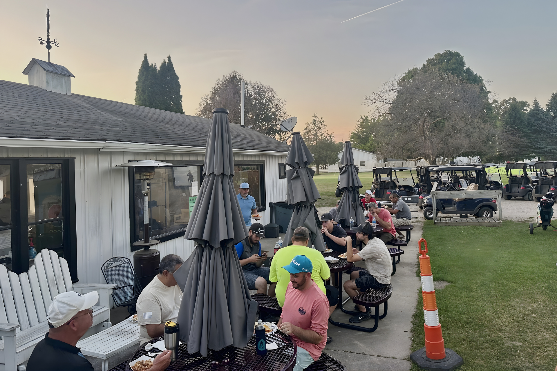 People enjoy an outdoor patio at a golf course, sitting at picnic tables with umbrellas near a white building at dusk.