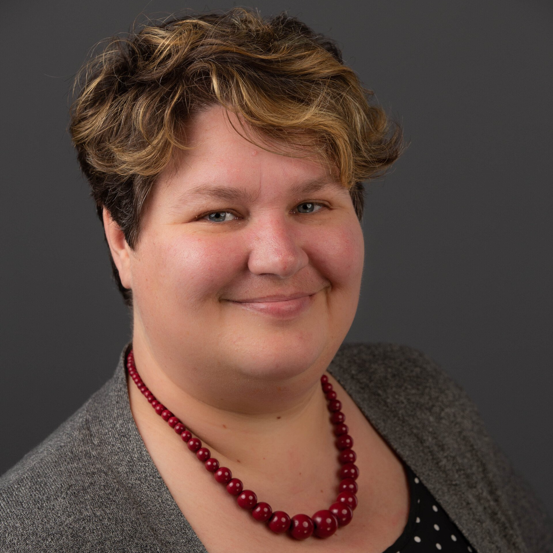 A woman wearing a gray jacket and a red necklace smiles for the camera