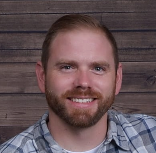 A man with a beard is smiling in front of a wooden wall