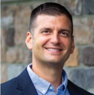 A man in a suit and blue shirt is smiling in front of a stone wall.