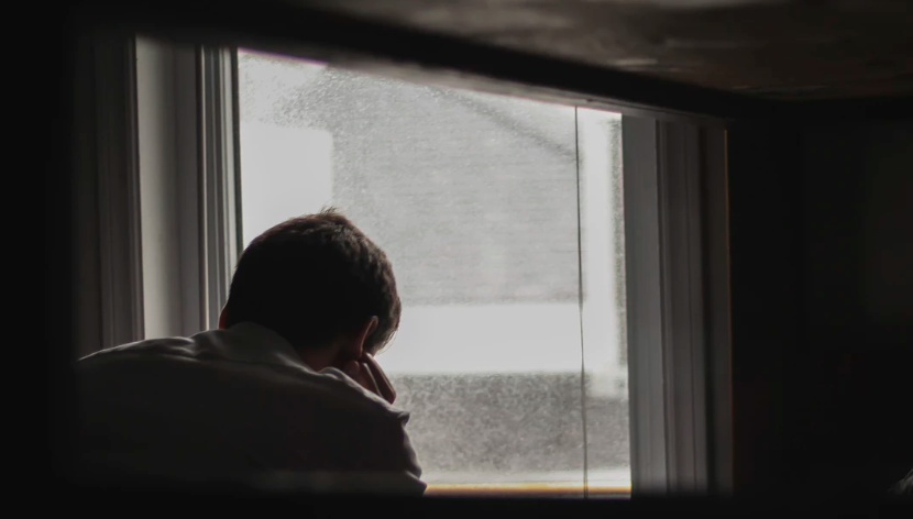 A man is sitting in front of a window looking out.