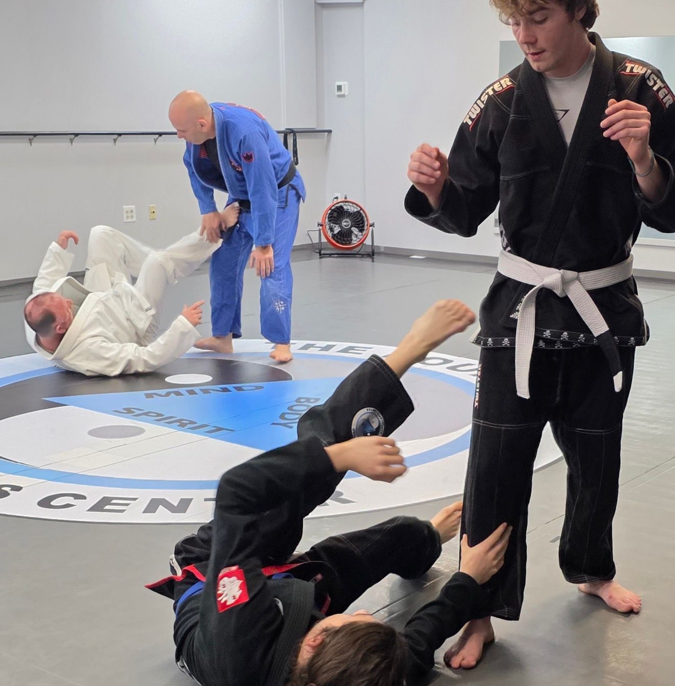 People practicing jiu-jitsu on mats, wearing gis in blue, black, and gray. Active grappling, gym setting.