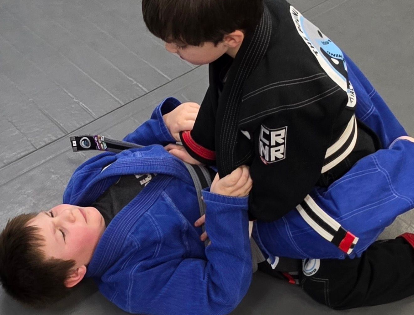 Two boys on a mat practicing Jiu-Jitsu. One boy is on top, giving a thumbs-up.