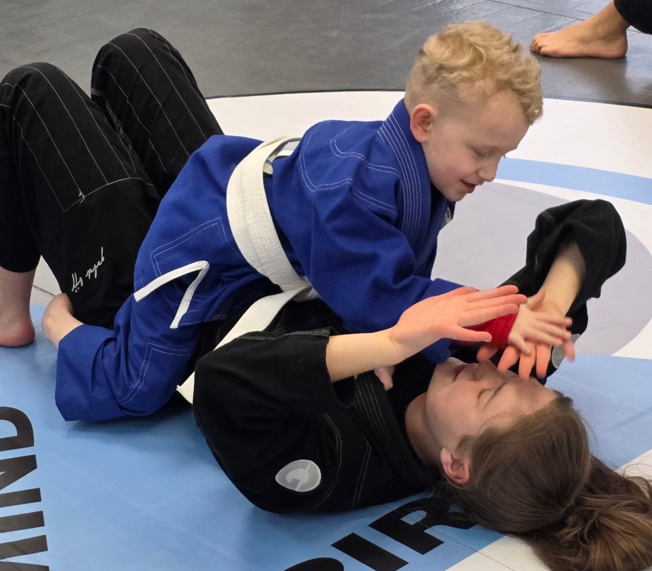 Two people grappling on a mat, one in a black gi, the other in a rash guard, legs entangled.