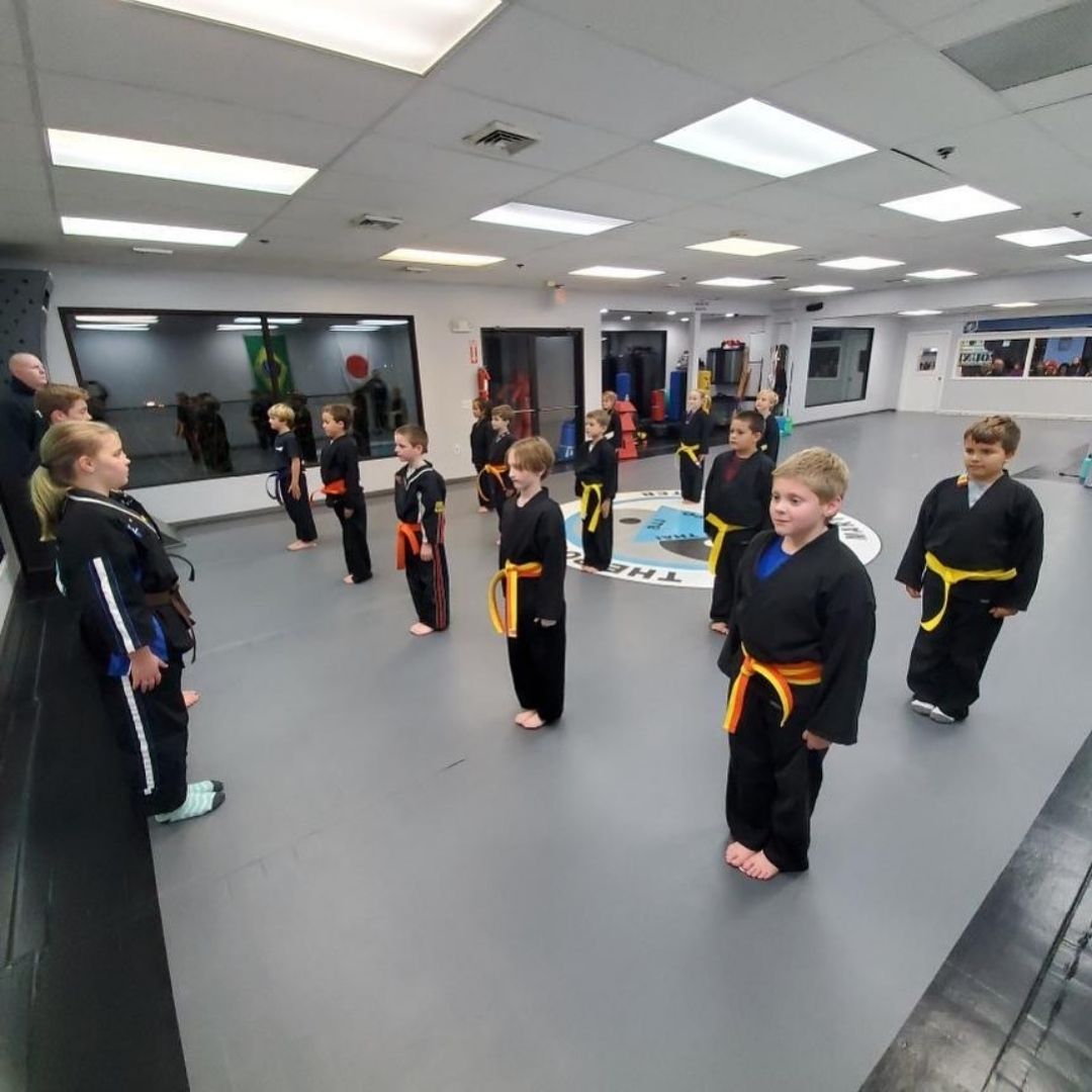 Children in colorful gis play a game on a mat. Two instructors pull a student in blue.