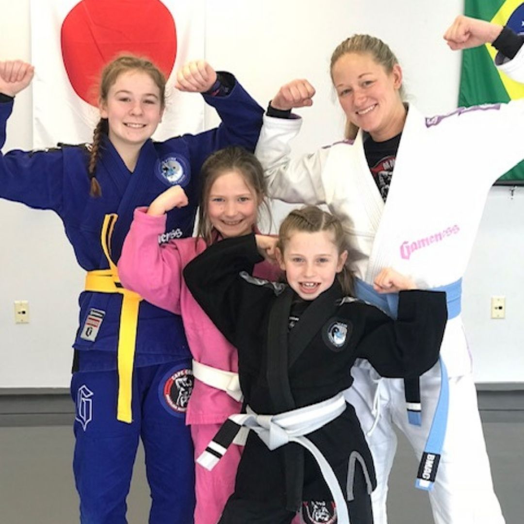Two young children in blue and black martial arts uniforms grappling on a gym floor, one smiling.