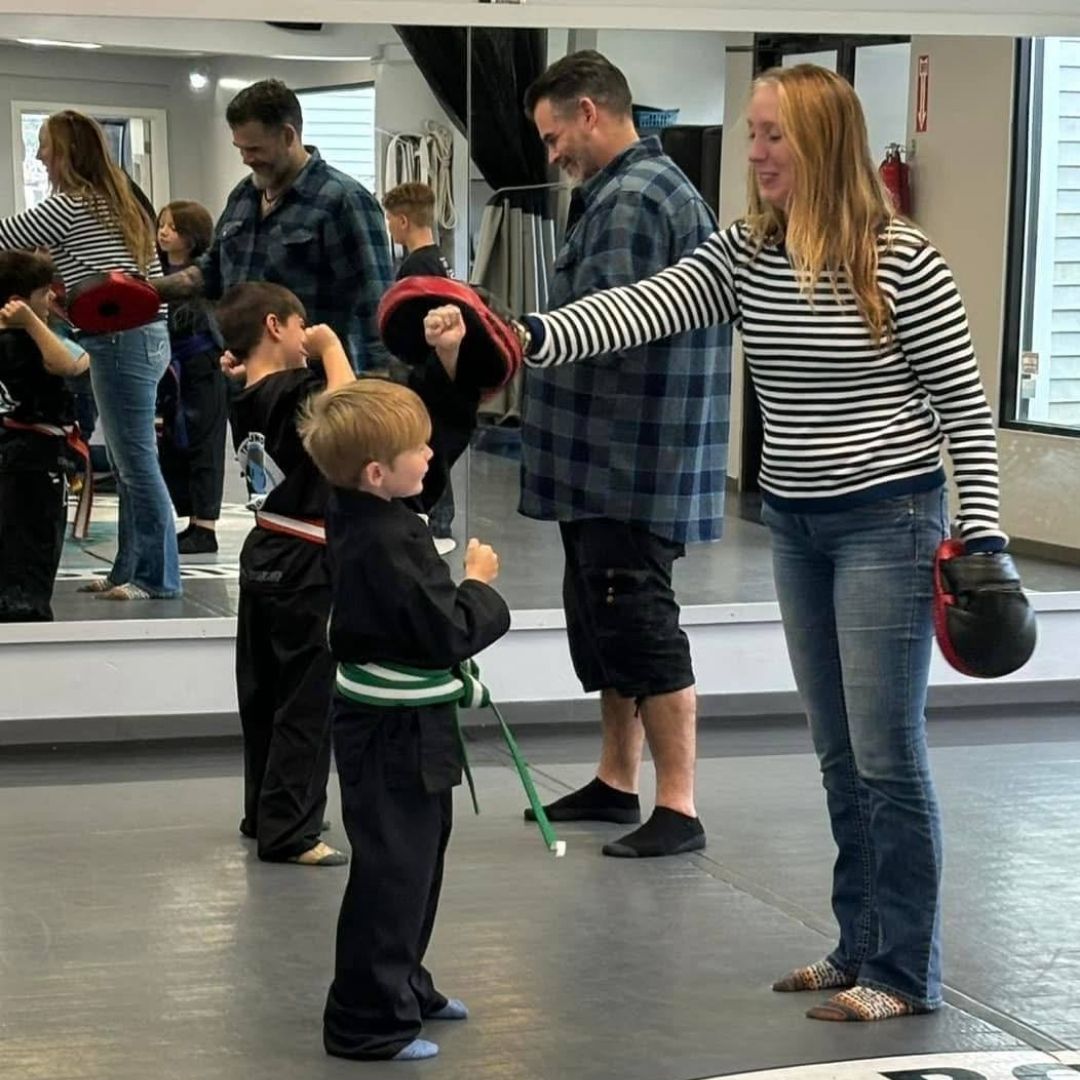 Two children in black and white martial arts uniforms practice a chokehold on a mat. One child smiles.