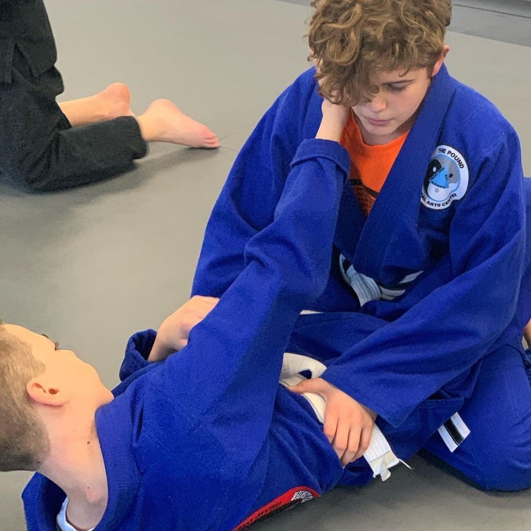Two children practicing Brazilian Jiu-Jitsu on a mat. One child in white gi has the other in a leg lock. A girl watches.