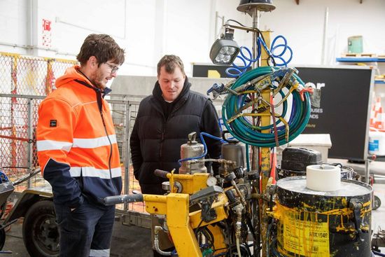Two men examining road marking equipment in a garage. One wears safety gear.