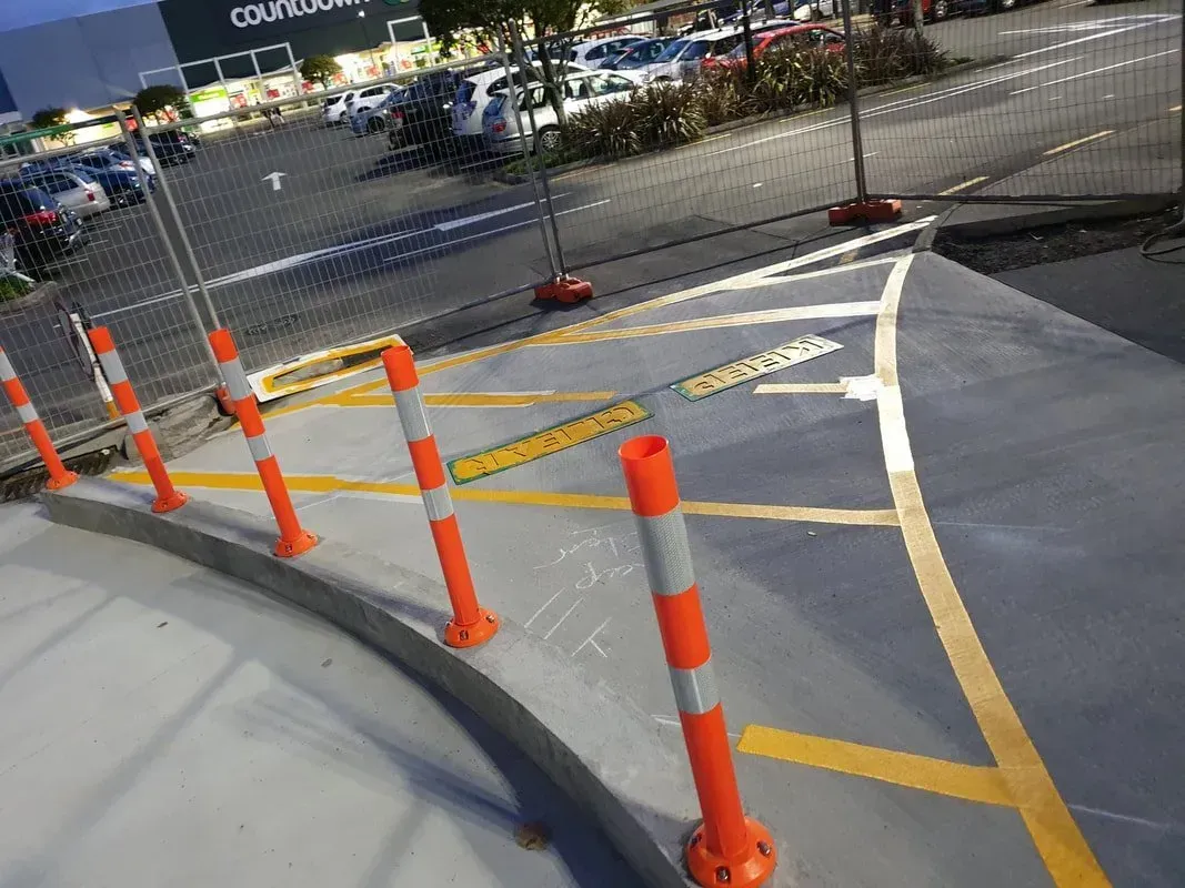 Orange and white bollards line a curved driveway. Yellow lines indicate parking space boundaries. A parking lot with cars is in the background.
