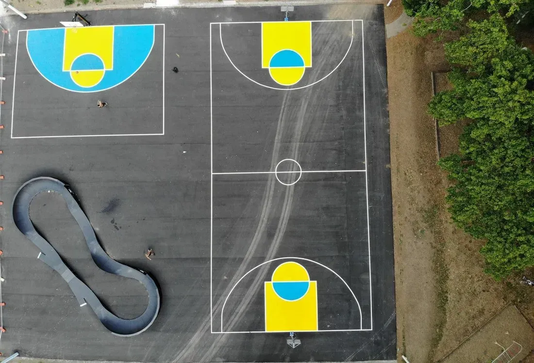 Aerial view of a paved sports court with a basketball court, half-court, and bike track; bright blue and yellow accents.