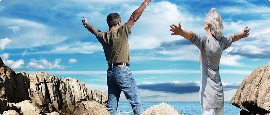 A man and a woman are standing on a rocky beach with their arms outstretched.