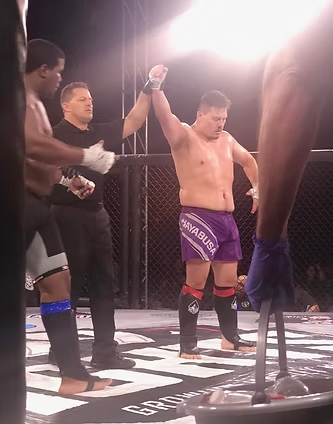 A man in purple shorts is standing in a boxing ring with his hand up.