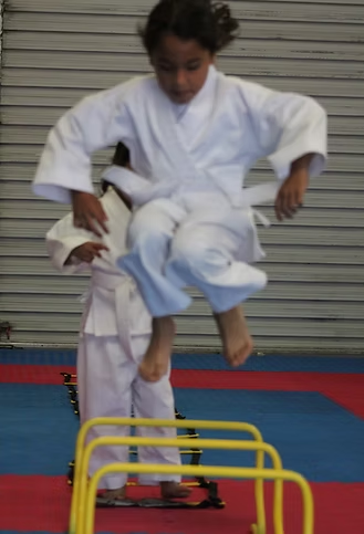 A girl in a white karate uniform is jumping over yellow hurdles