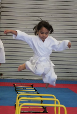 A little girl in a white karate uniform is jumping over hurdles