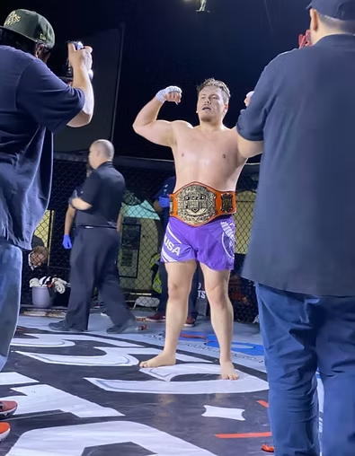 A man in purple shorts is standing on a boxing ring with a belt around his waist.