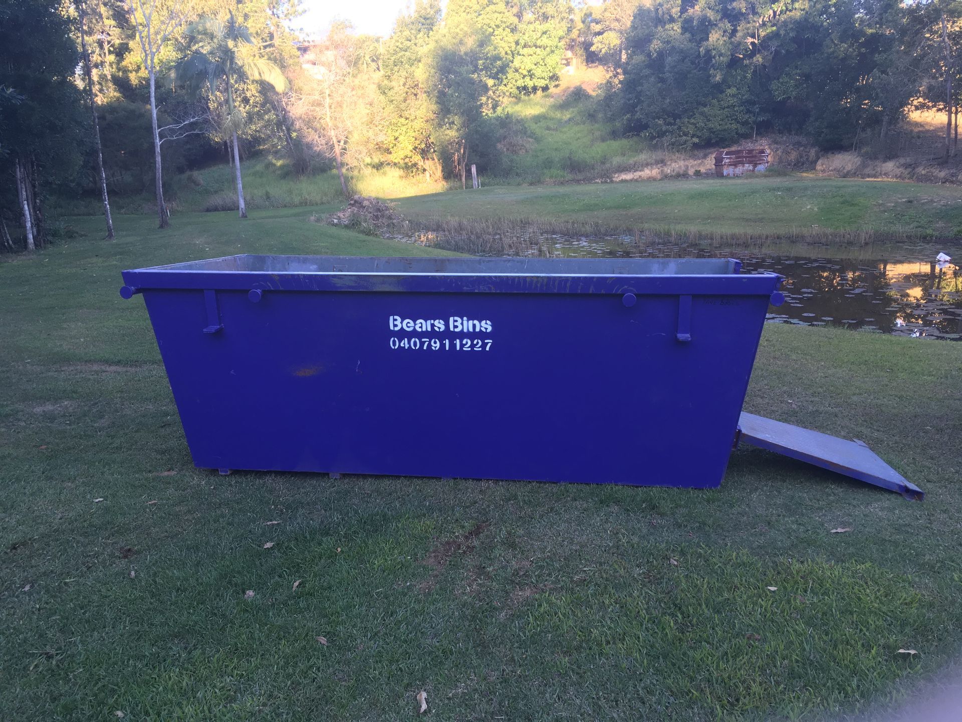 Blue dumpster on grass, near a pond with trees in the background — Bears Bins Skip Hire In Meridan Plains, QLD