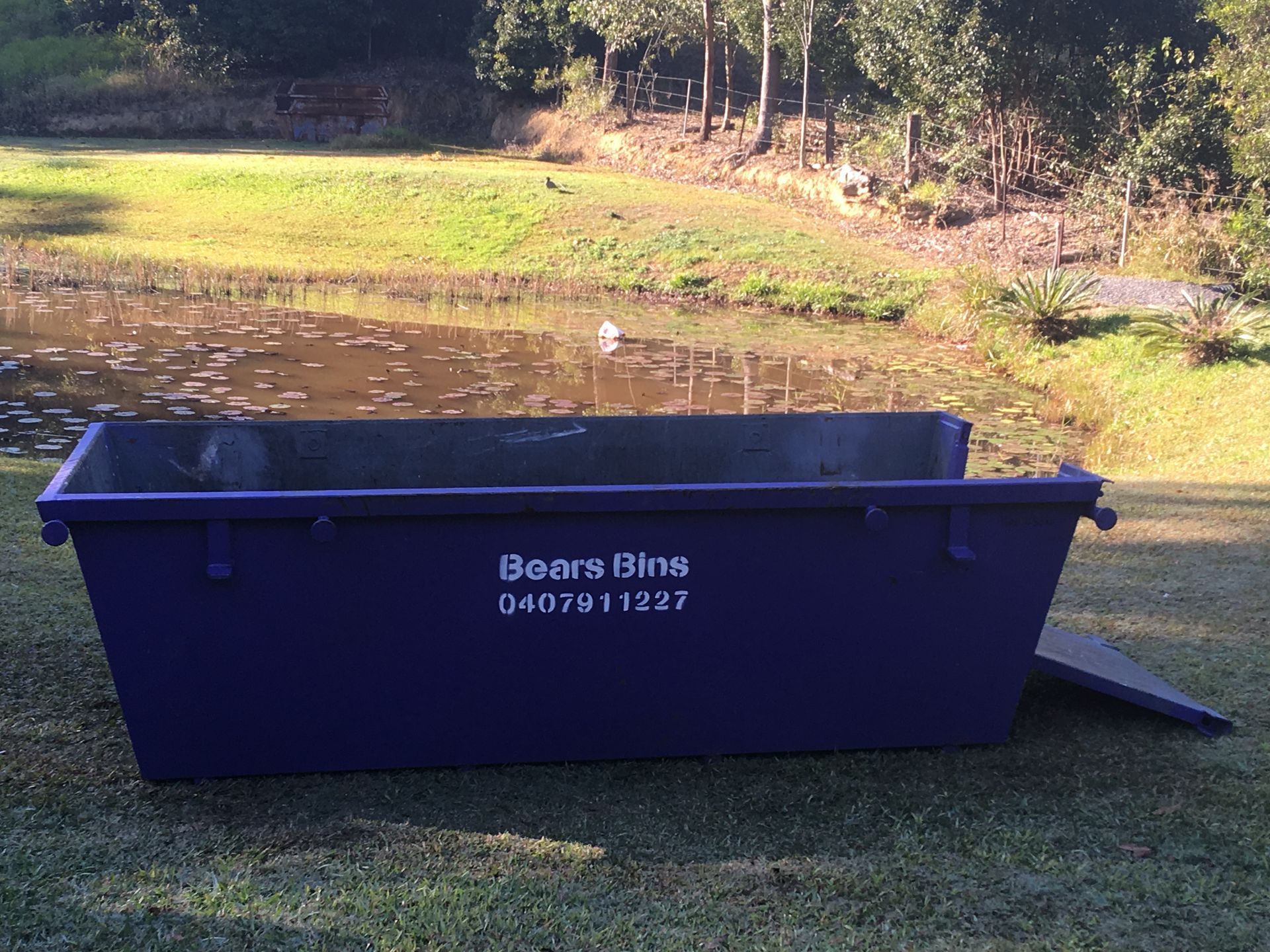 Blue skip bin on grass near water with 
