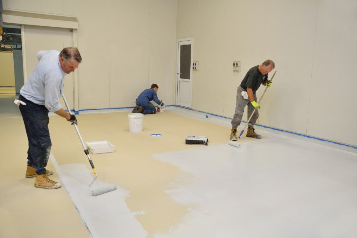 Three people painting a floor white in an indoor room with blue tape along the edges.