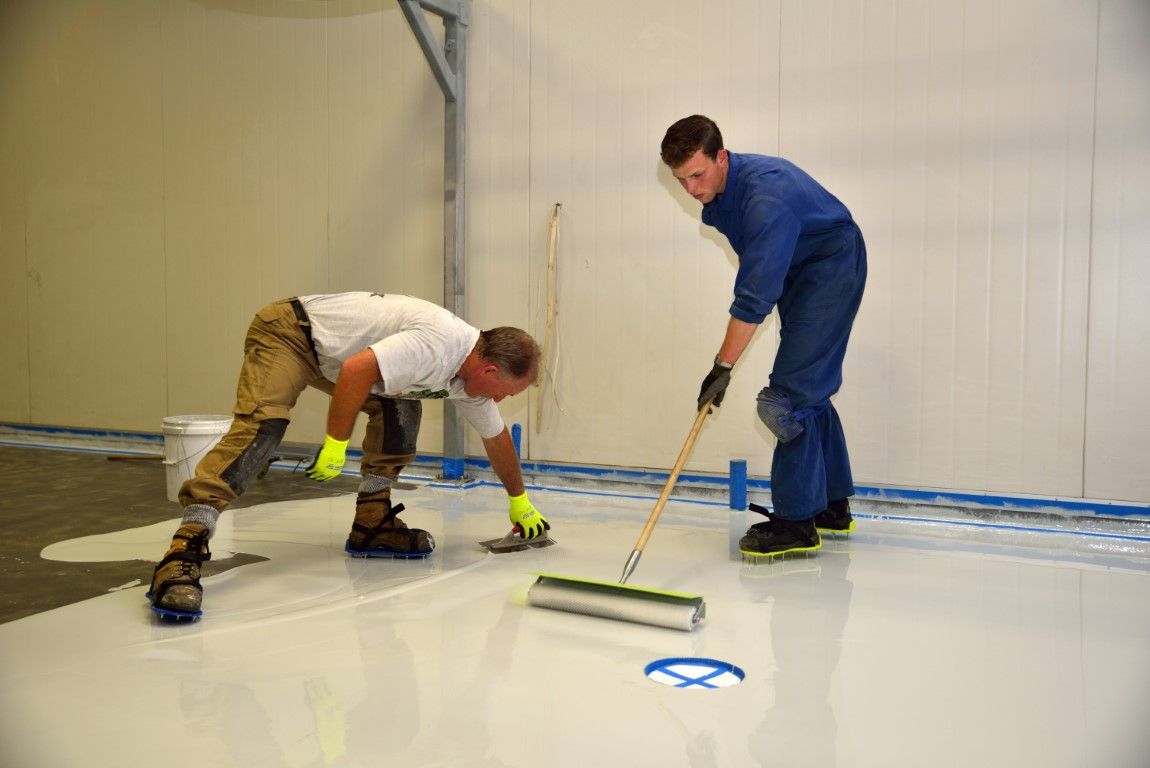 Two workers applying epoxy flooring to a large, white-walled room; one kneels, the other uses a roller.