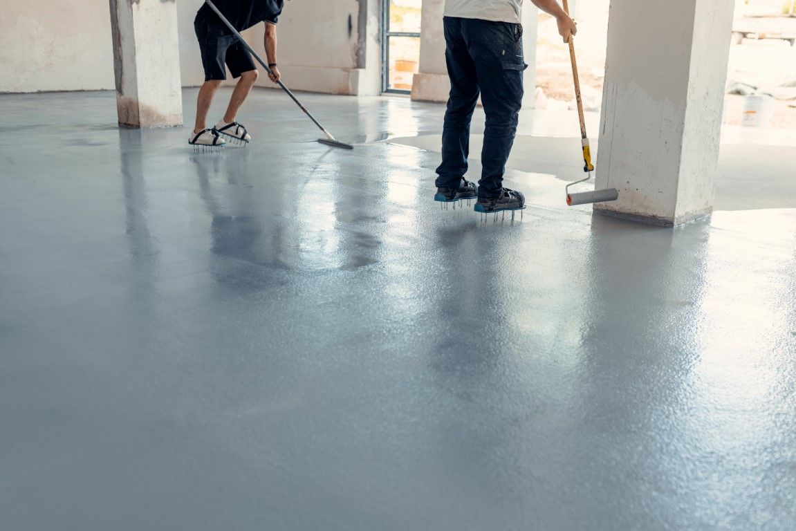 Workers applying gray epoxy coating to a concrete floor in an unfinished building.