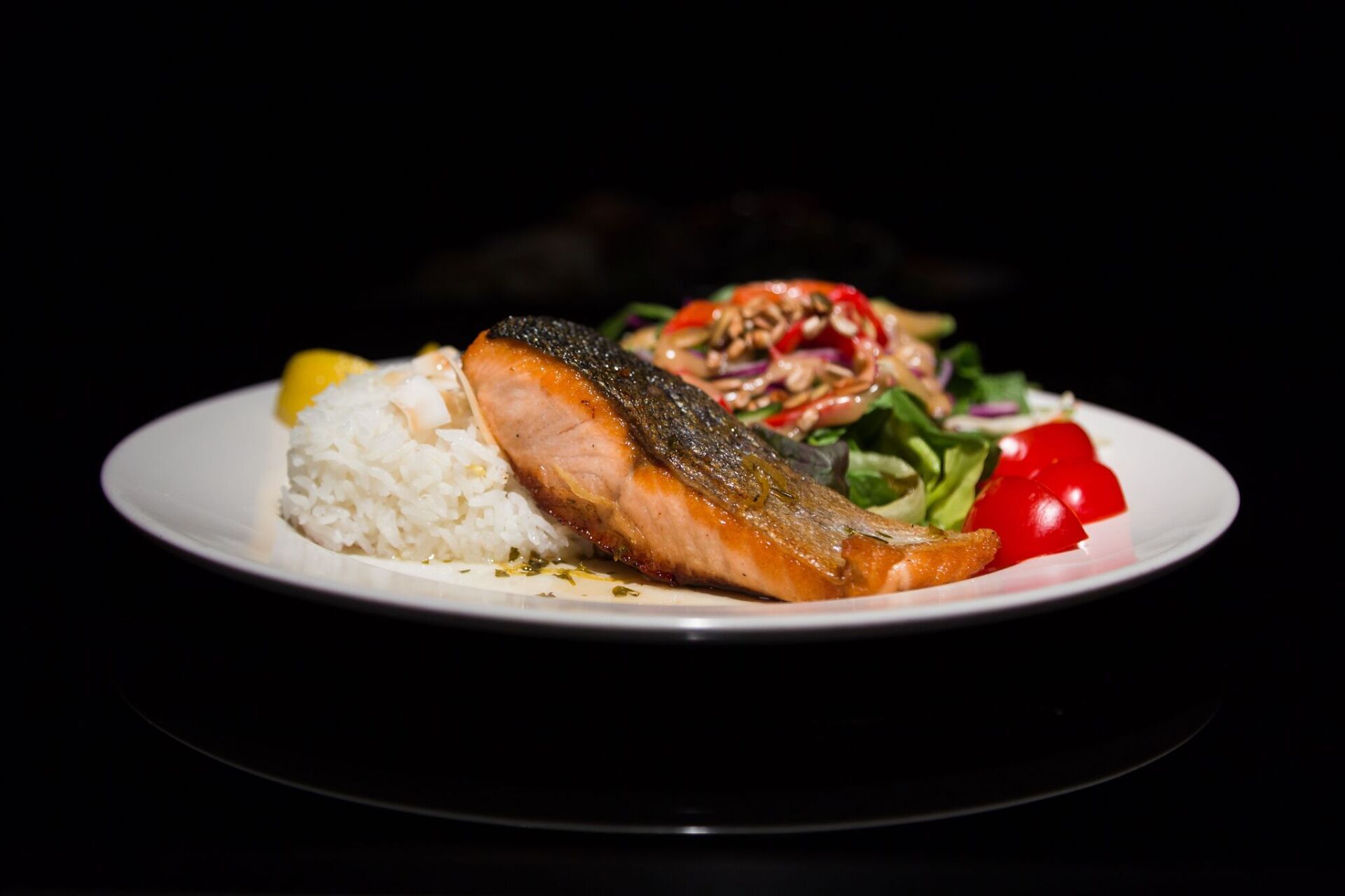 A white plate topped with salmon , rice and vegetables on a black background.