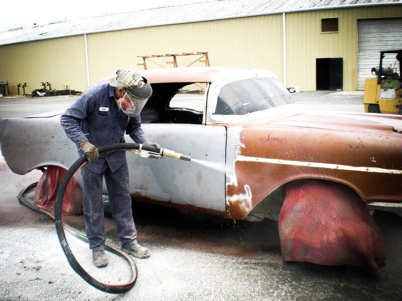 A man wearing a mask is sandblasting an old car