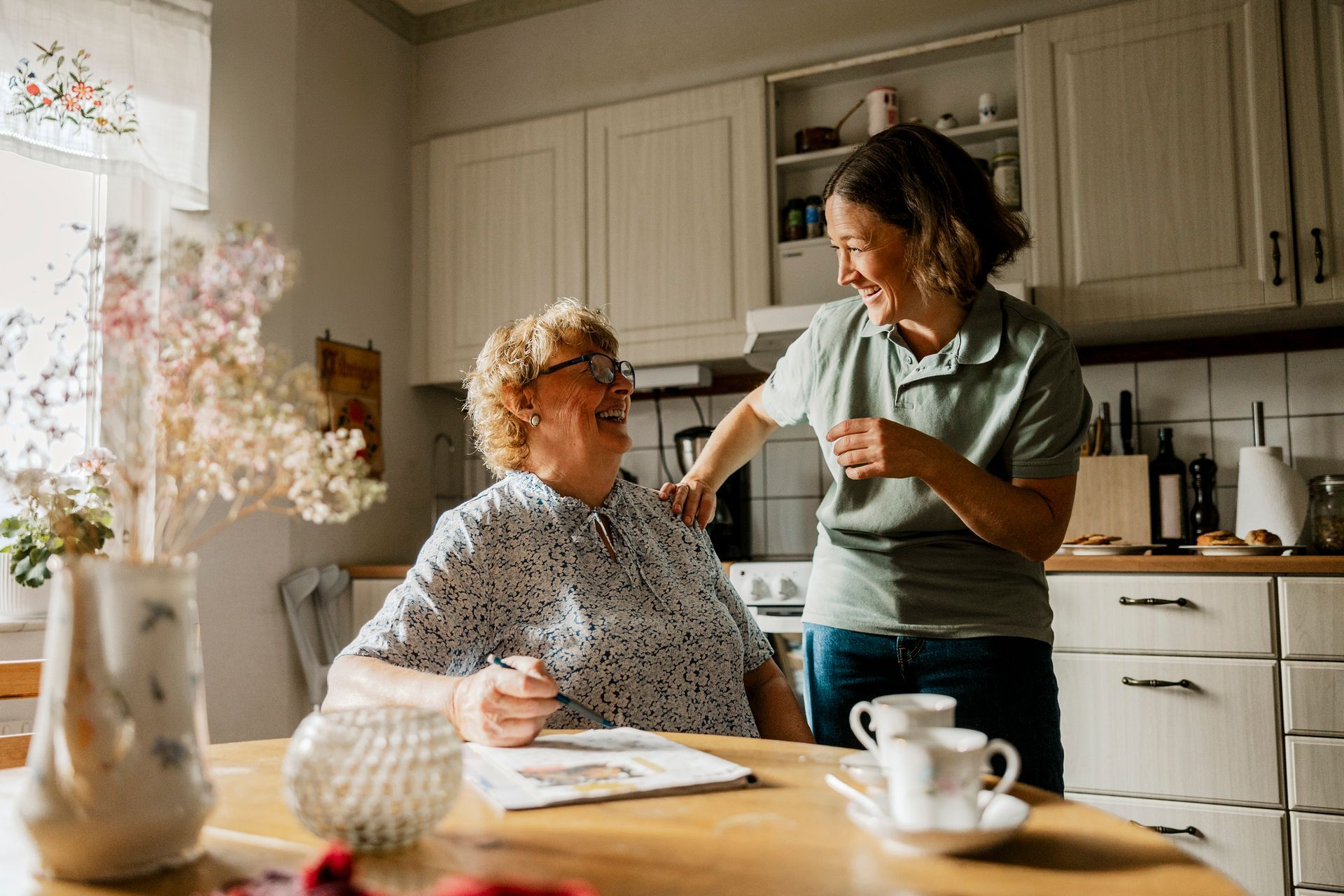 Senior woman receives assistance at kitchen table with flowers, papers, and warm home setting.