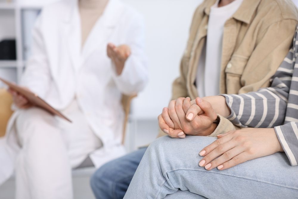Doctor speaking with a seated patient in a clinic, with hands clasped in the foreground.