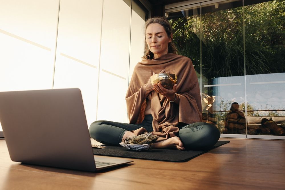 Woman in meditation pose with laptop, holding a bowl. She is wrapped in a shawl, in a sunlit room.