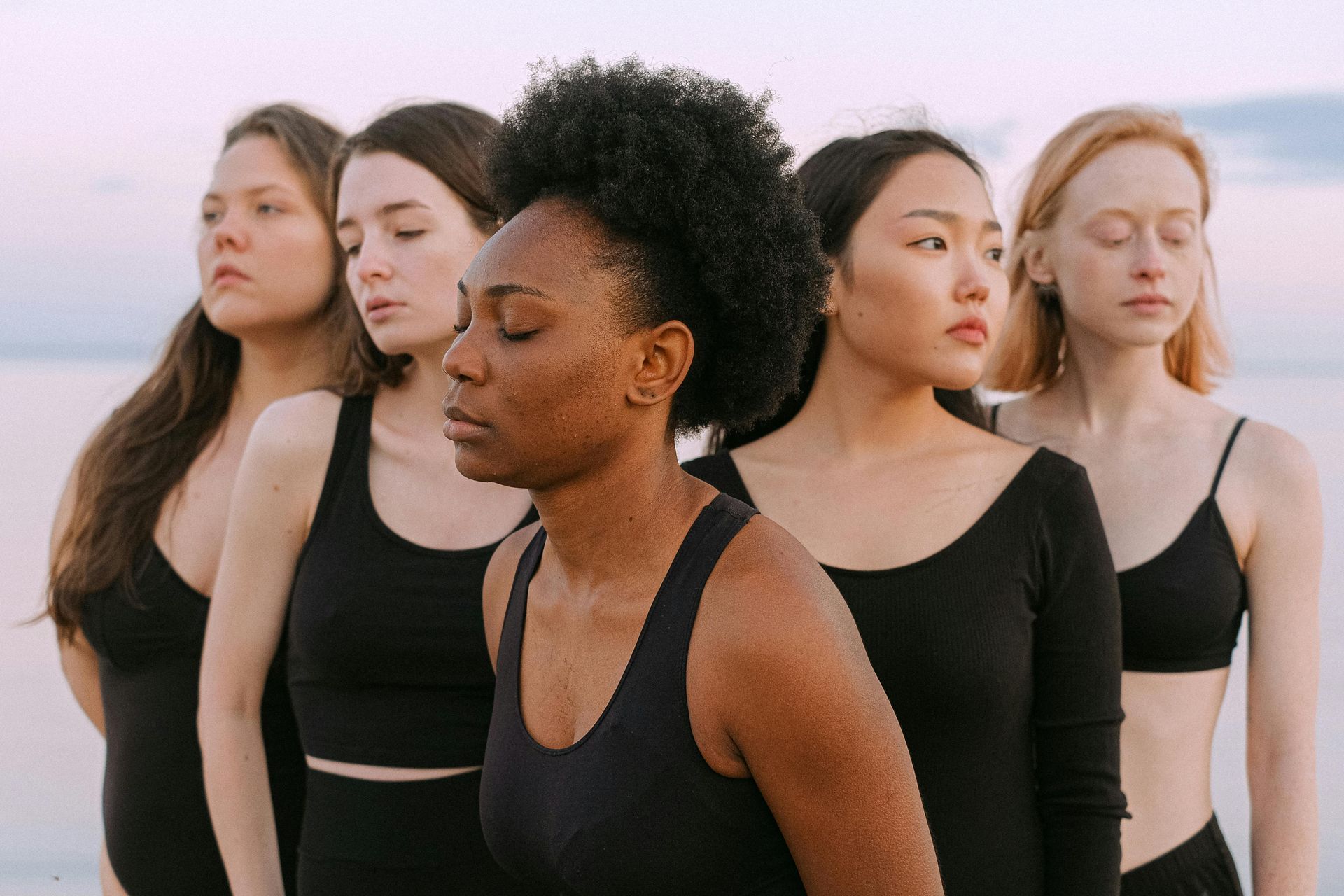 Five racially, ethnically diverse women standing outside dressed in various types of black leotards