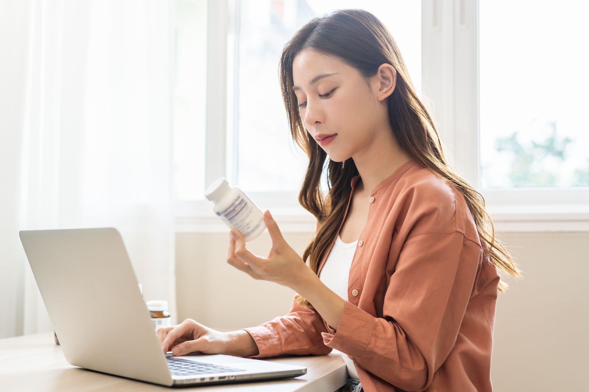 A woman is holding a bottle of folic acid while using a laptop computer to research folic acid supplements for fertility