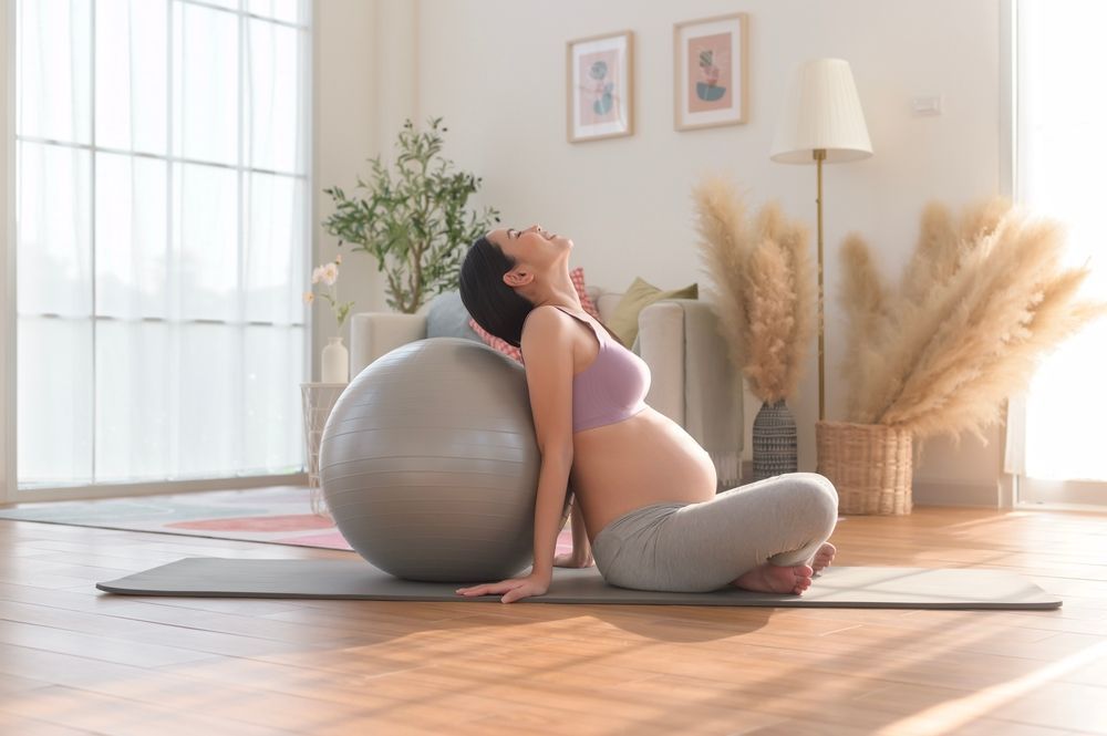 A pregnant woman is sitting on a yoga mat with an exercise ball.