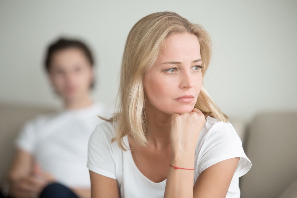 A woman is sitting on a couch with her hand on her chin while a man sits behind her.