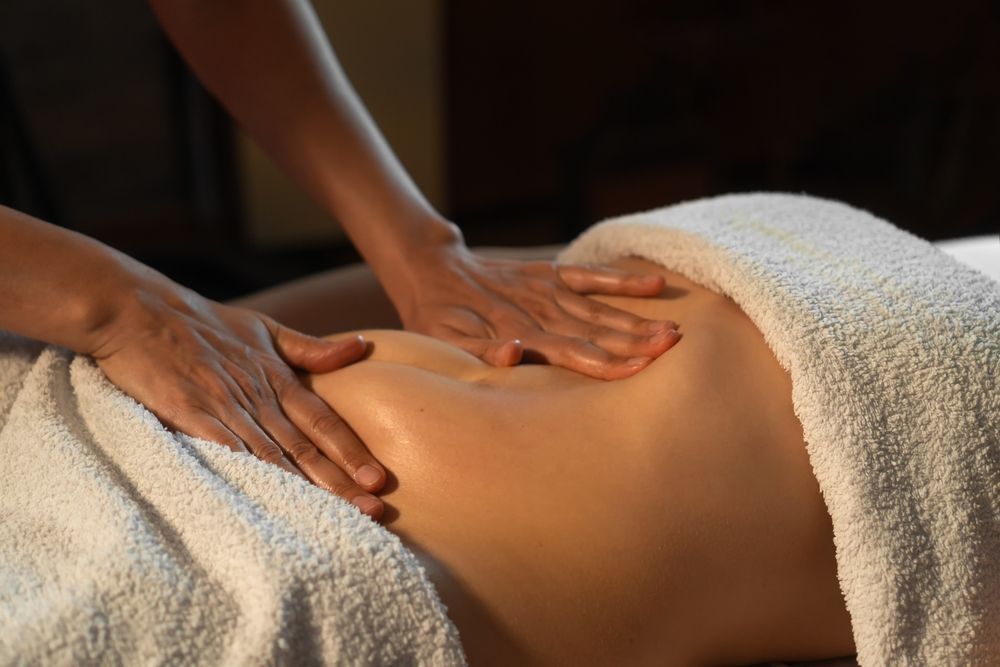 Hands performing a gentle abdominal massage on a person lying down under a white towel.