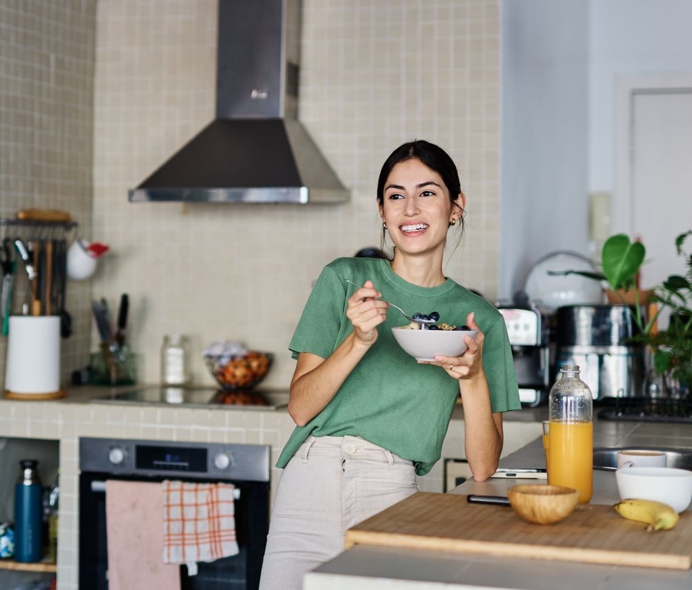 Woman smiling in a kitchen, holding a bowl and spoon beside a table with orange juice and fruit.