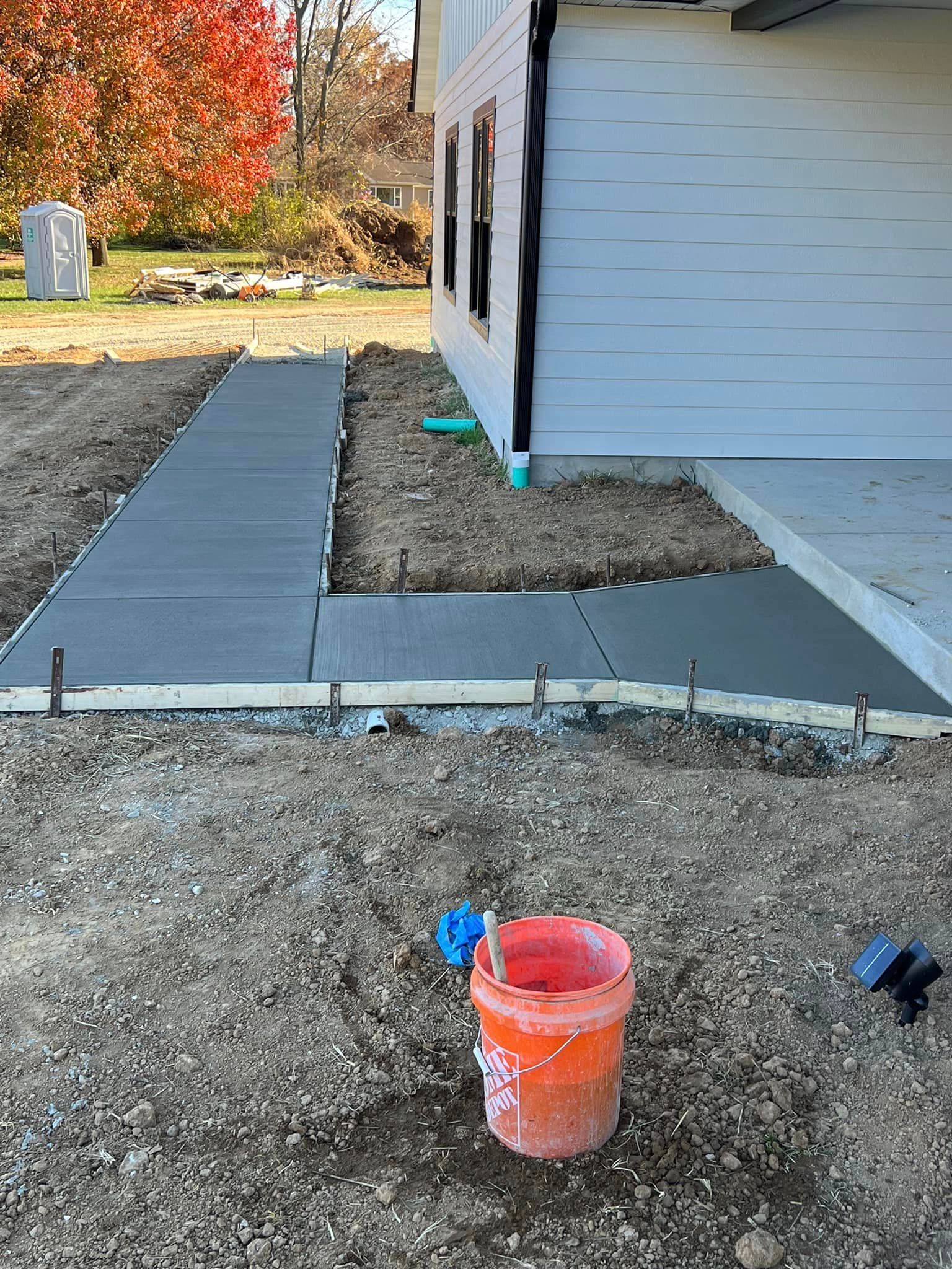 A bucket of concrete is sitting in the dirt in front of a house.