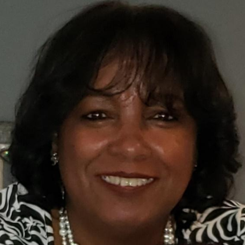 Belinda Eleby with short dark hair, smiling, wearing a pearl necklace and black and white patterned top.