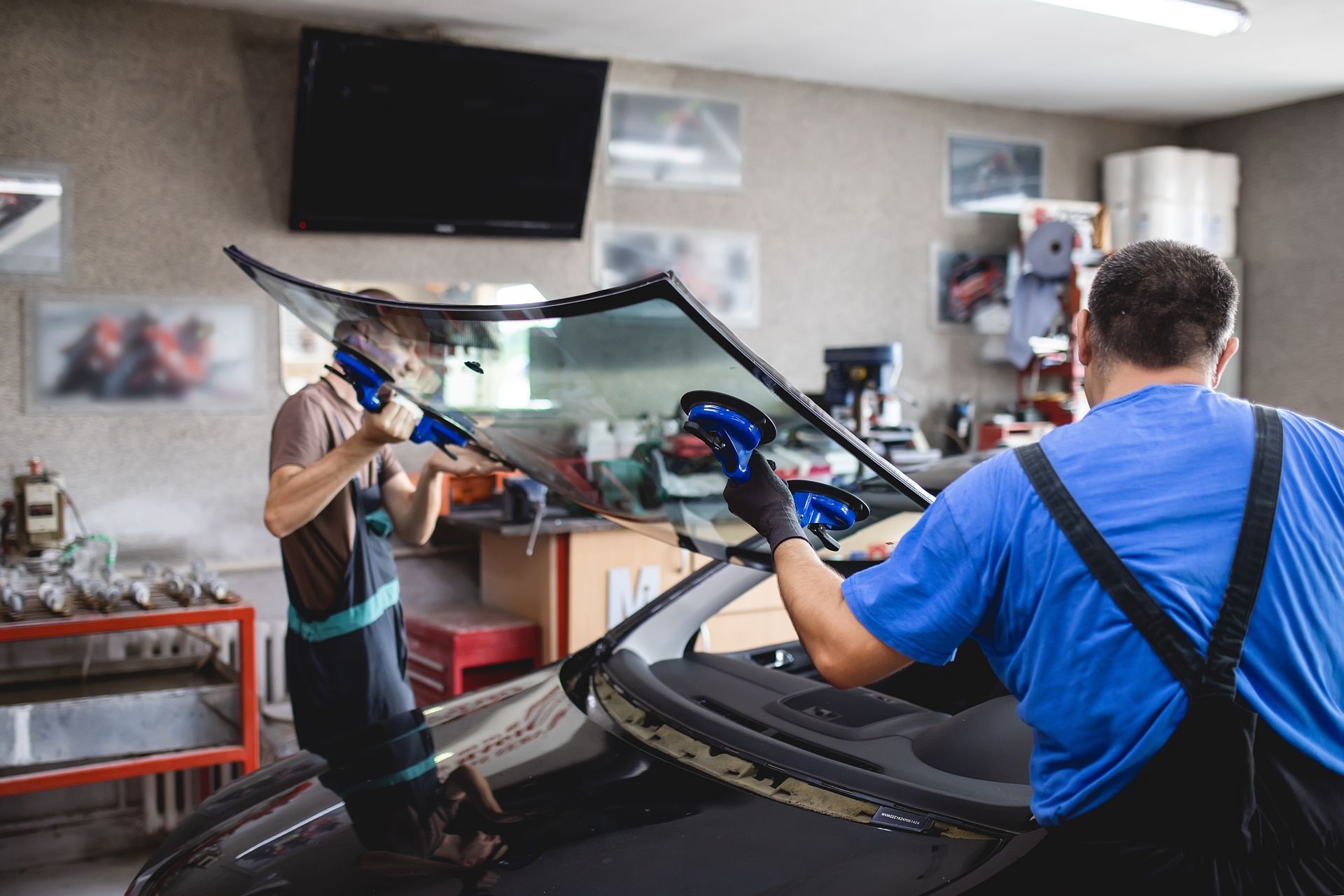 Two auto repair technicians using blue suction cups to lift a car windshield in a garage.