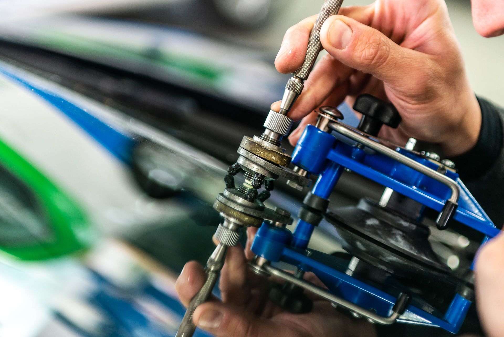 Two auto repair technicians using blue suction cups to lift a car windshield in a garage.
