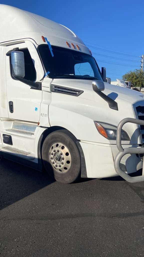 White semi-truck parked on asphalt road under a clear, blue sky. The truck has a metal bumper.