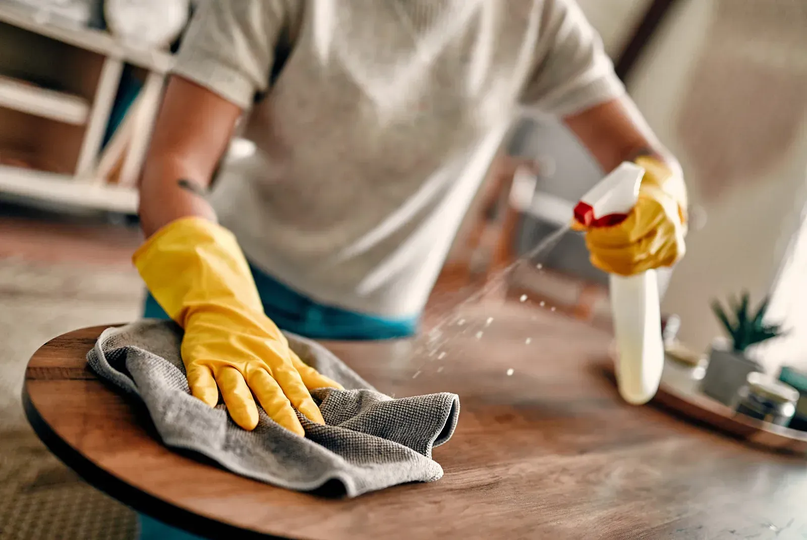 A person wearing yellow gloves is cleaning a wooden table with a cloth and spray bottle.