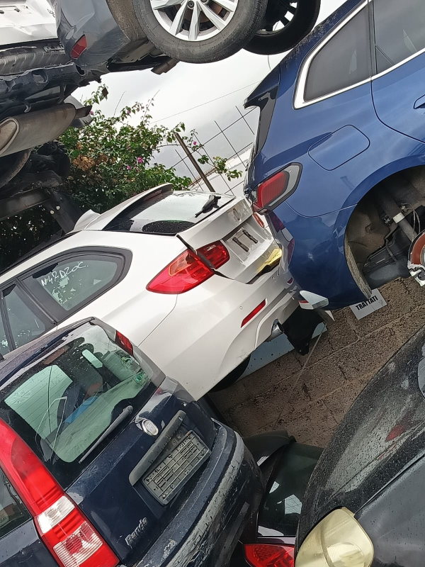Un deposito di rottami ingombro di auto presenta diverse vetture accatastate e parcheggiate, tra cui un SUV bianco e un veicolo blu scuro.