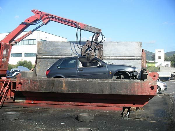 Sotto un cielo azzurro, un'enorme gru meccanica solleva un'auto grigio scuro all'interno di un deposito di rottami metallici.