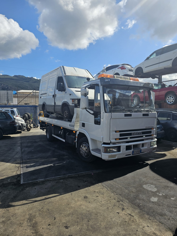 Un carro attrezzi Iveco bianco che trasporta un furgone bianco in un deposito veicoli all'aperto sotto un cielo azzurro.