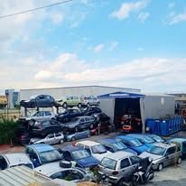 Un deposito di rottami con auto accatastate e parcheggiate all'esterno di un edificio industriale, sotto un cielo azzurro.