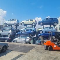 In un deposito di rottami all'aperto, diverse auto sono accatastate in fila, con un carrello elevatore arancione parcheggiato in primo piano.