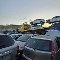 Un carro attrezzi giallo che trasporta un'auto bianca in uno sfasciacarrozze pieno di veicoli danneggiati, sotto un cielo sereno.